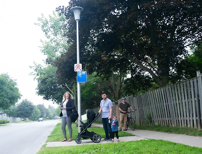 Photo of people waiting for the bus at a stop with the blue bus plate on the pole.